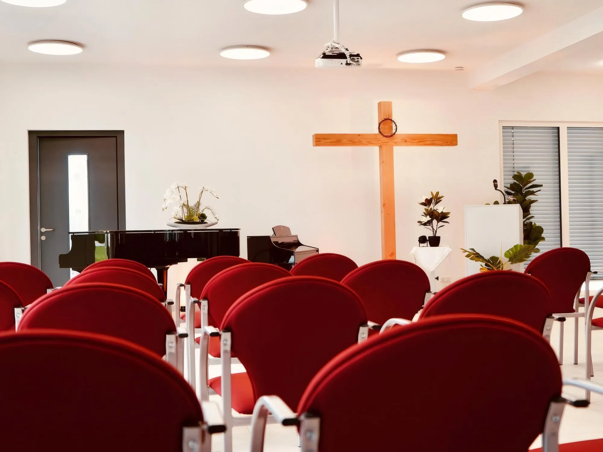Heller Gemeindesaal mit Holzkreuz, Rednerpult und roter Bestuhlung