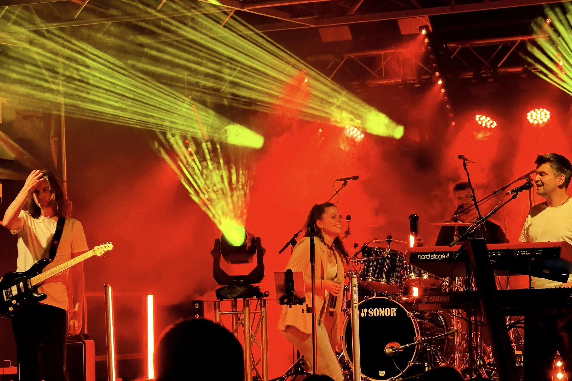 Liveband auf der Bühne, rot ausgeleuchtet, mit gelben Laserstrahlen über dem Publikum