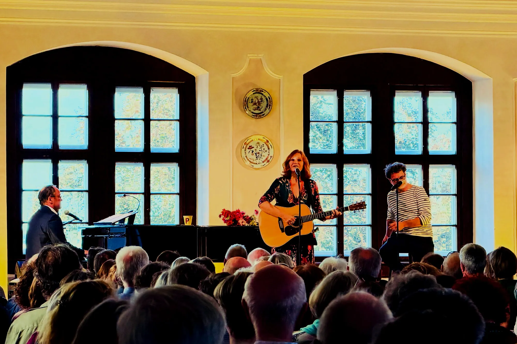 Konzert im Schlosssaal: Sängerin mit Gitarre, Pianist am Flügel und Perkussionist vor vollbesetzten Stuhlreihen